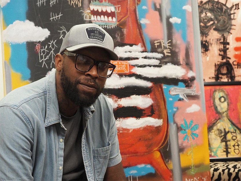 A Black man with a beard wearing a gray baseball cap, black glasses, a dark gray T-shirt, and a short-sleeved denim shirt poses in front of a colorful artwork