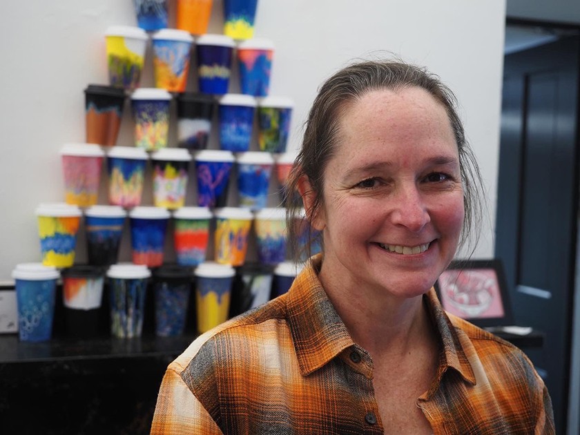 A white woman with pulled-back brown hair and a orange, brown, and white collared shirt smiles and poses in front of a display of colorful tumblers