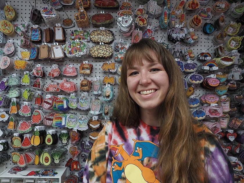A white woman with long brown hair and bangs wears a colorful tie-dye shirt with a cartoon flying dragon on it. She's smiling and posed in front of a wall displaying colorful stickers