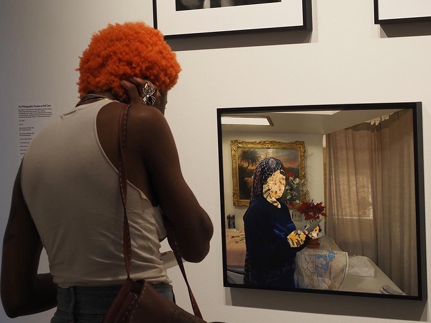 A person stands with their back to the camera. They're looking at framed artwork hanging on a gallery wall. The photograph in front of them shows a woman with their face and hands Photoshopped as floral patterns.
