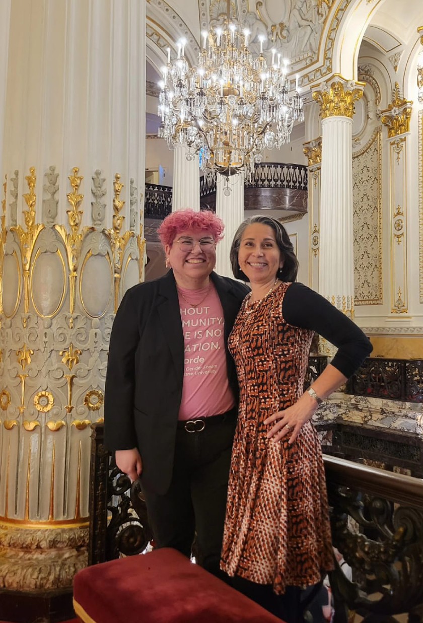 Two Latine people pose smiling for the camera. The person on the left has short, curly pink hair and is wearing a pink T-shirt, dark slacks, and a Black jacket. The person on the right has shoulder-length straight dark hair and is wearing a brown patterned dress with black sleeves 