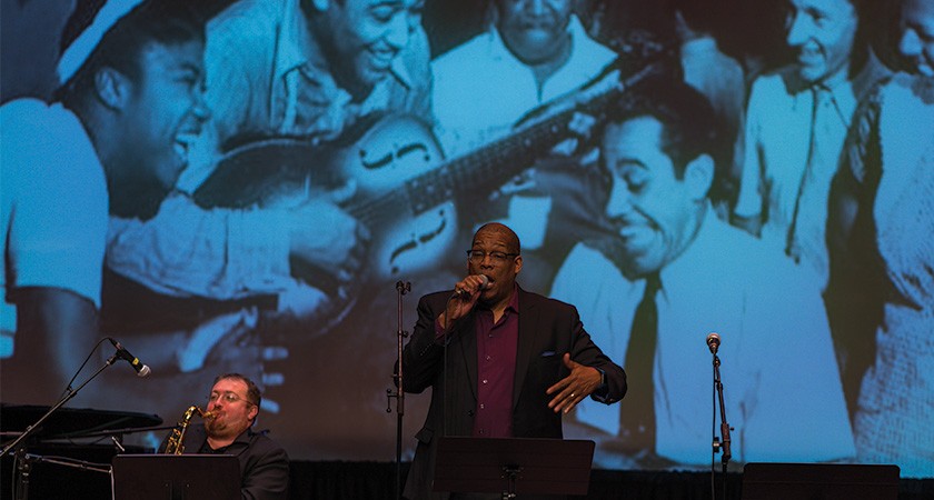 A person in a burgundy shirt and a black blazer holds a microphone in front of a large sepia-toned photograph of smiling people surrounding a guitar player 
