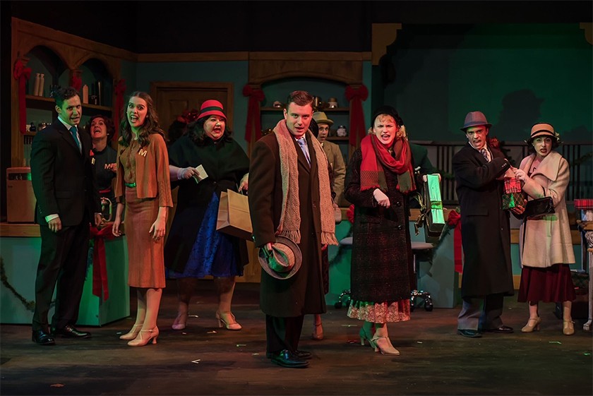 A group of nine actors who contain a mix of genders and ages stand in a row on stage. Some are wearing hats and long overcoats and scarfs. All have their mouths open and appear to be singing angrily. 
