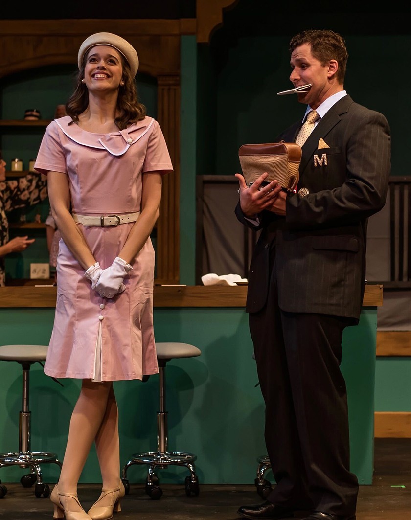A woman in a pink dress, tan hat, and white gloves stands while smiling broadly. Beside her, a man in a dark brown suit looks past her while holding a piece of paper in his mouth and a bag in his hands.