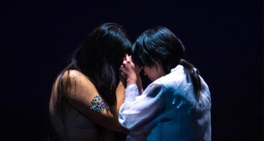 Two people with long straight black hair stand extremely close to one another, pressing their faces together