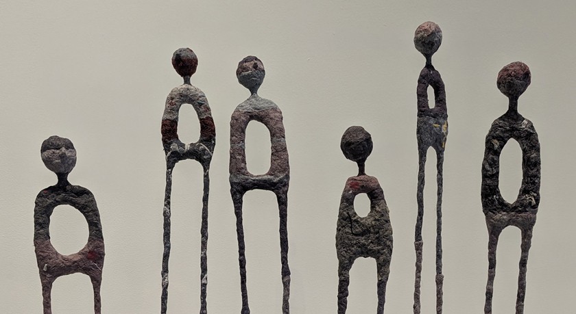 A close-up of six skinny sculptures created from dryer lint. The sculptures are similar to human form, showing long legs, a torso with missing arms and a hole in the stomach, and a faceless head