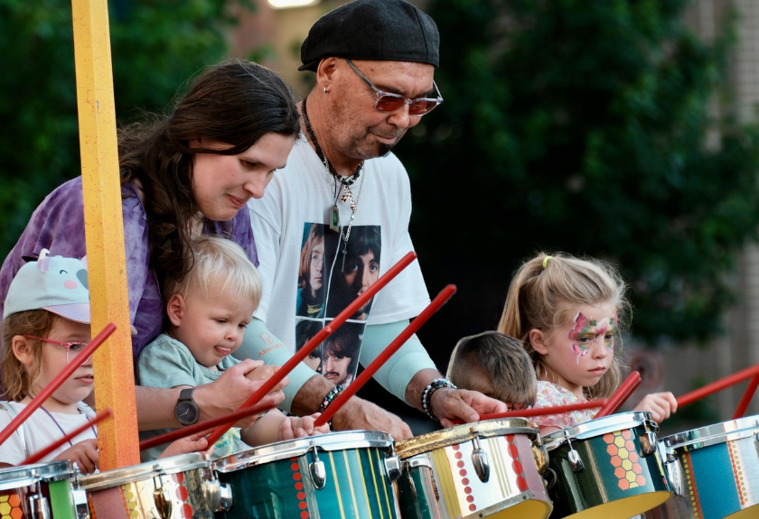 A man and woman help three young children play drums during an outdoor event.