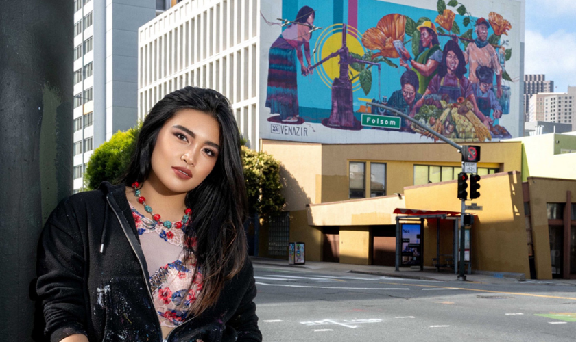 A person with long dark hair stands on a city street corner with a colorful mural of people and nature painted on a building in the background.