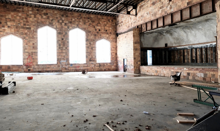 An empty, abandoned warehouse with large arched windows, exposed brick walls, scattered debris, graffiti, and a single office chair facing a raised platform area.