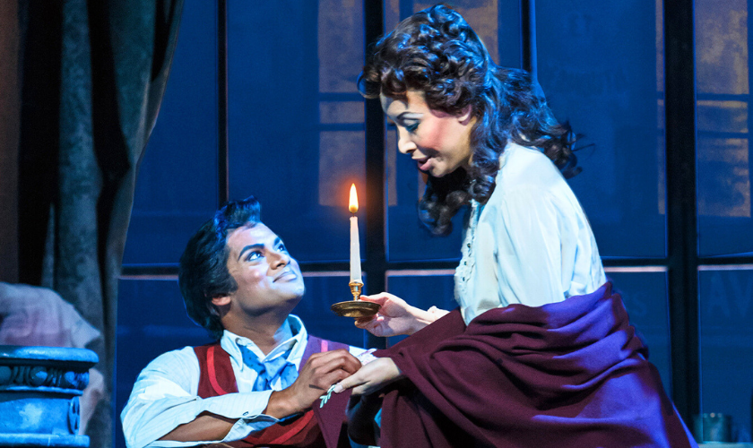 Two opera performers in period costumes are on stage; a man kneels, holding a woman’s hand as she holds a lit candle. They appear to be in an emotional scene, with dramatic lighting and a window in the background.