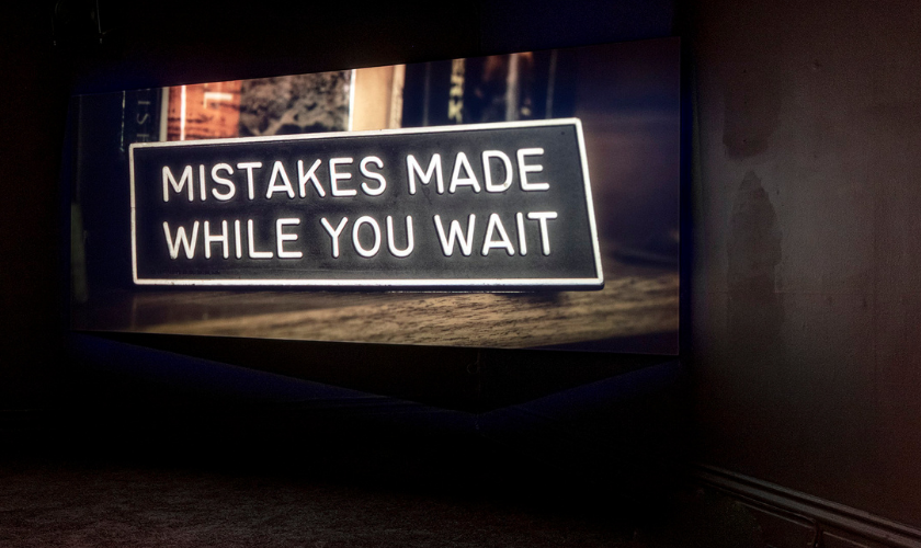 A sign with white text on a black background reads, MISTAKES MADE WHILE YOU WAIT, displayed on a screen in a dimly lit room.