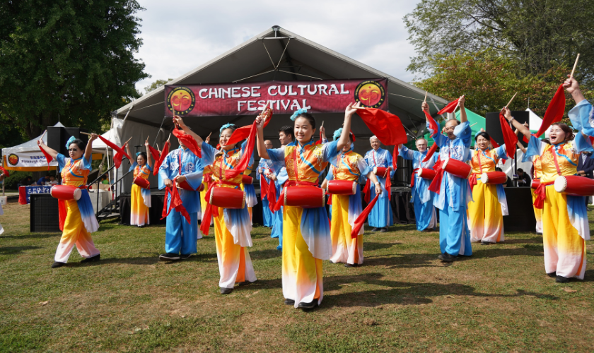 A group of performers in colorful traditional Chinese costumes play red drums and wave red cloths on a grassy outdoor stage under a tent with a “Chinese Cultural Festival” banner. Spectators and trees are visible in the background.