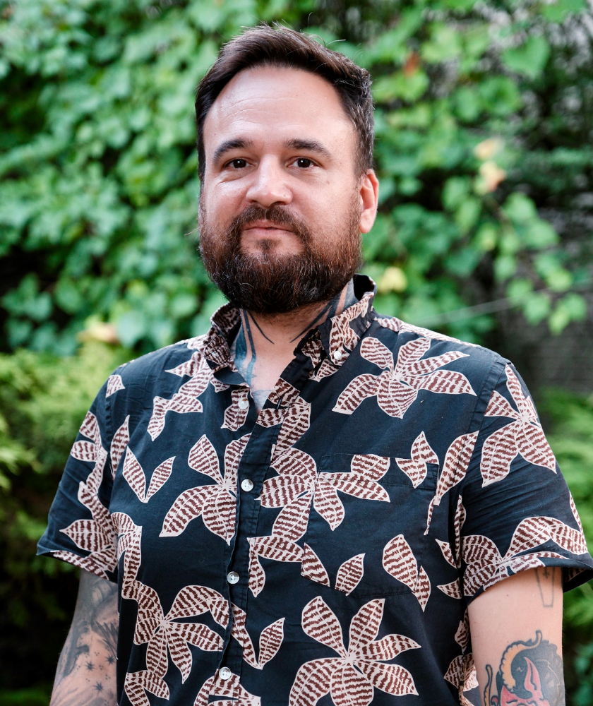 A bearded man with dark hair stands outdoors in front of greenery, wearing a black shirt with a bold, light-colored leaf pattern. He has tattoos on his arms and looks at the camera with a slight smile.