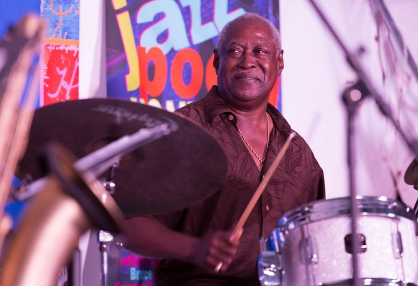 Smiling man playing drums at a jazz event, colorful jazz poster in background.