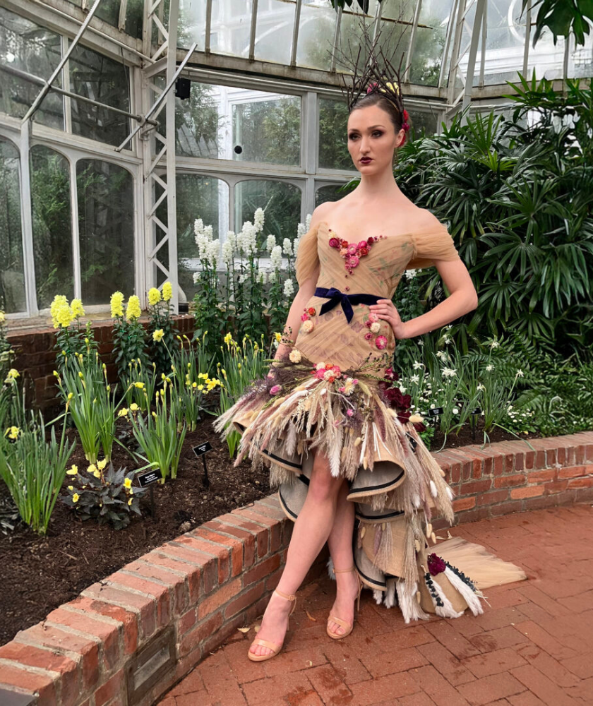 A person poses in a greenhouse wearing an elaborate beige dress decorated with flowers and plants, with a dark ribbon at the waist. They stand by a garden bed with blooming flowers and lush greenery in the background.