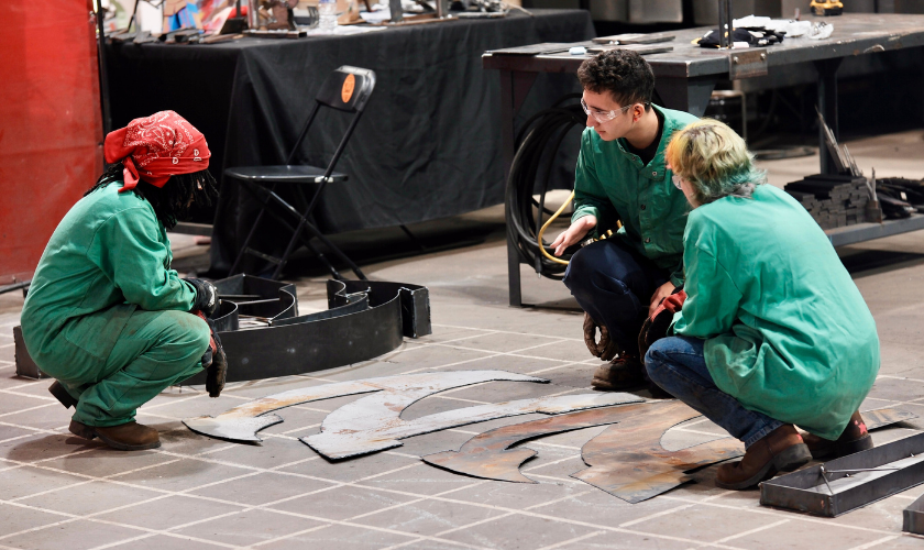 Three people wearing green protective jackets and gloves crouch on a workshop floor, examining large, irregularly shaped metal pieces laid out in front of them. Tools and equipment are visible in the background.