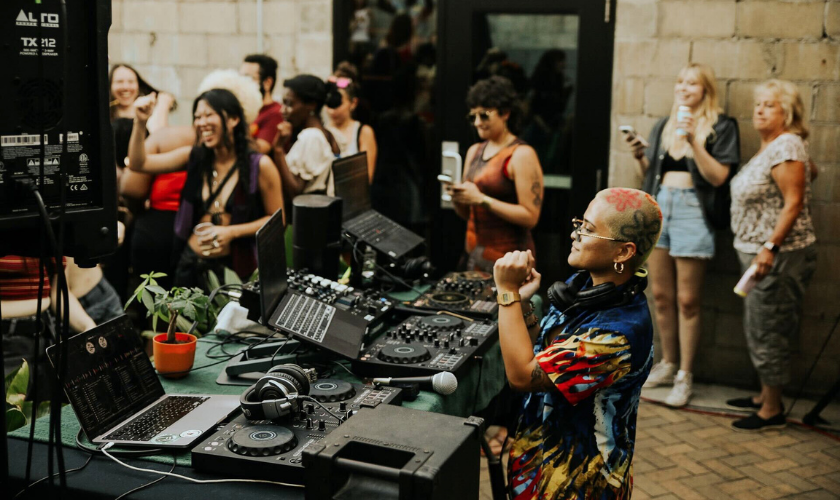 A DJ with tattoos and glasses performs behind turntables and computers as people stand, watch, and enjoy the music in an indoor venue with brick walls.