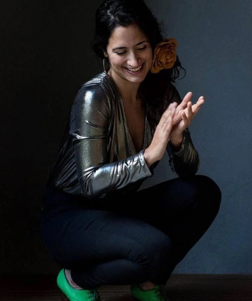 A woman with long dark hair, wearing a metallic silver top, black pants, and green shoes, crouches and smiles while clapping her hands. She has a large yellow flower in her hair against a dark background.
