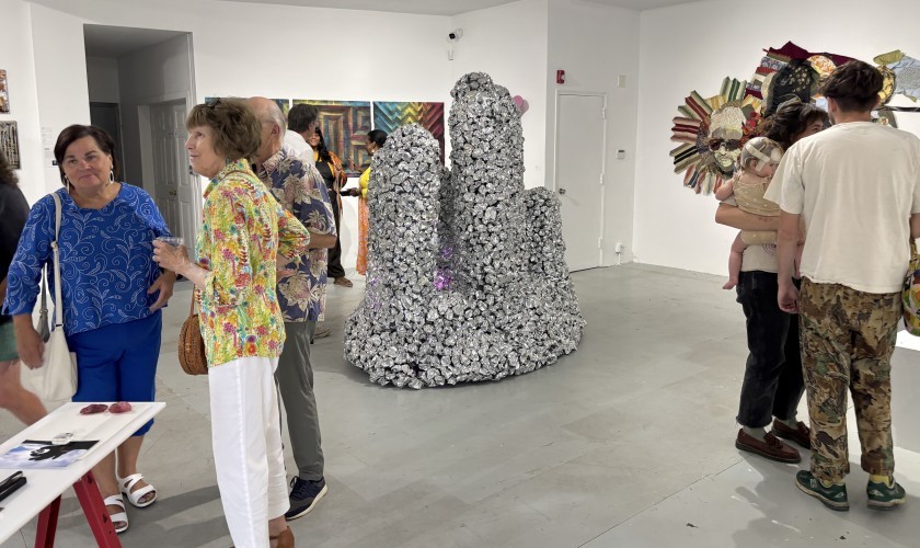 People mingle in a contemporary art gallery with sculptures and colorful wall art. A central silver cluster sculpture stands out, while visitors, including a person holding a child, observe the artwork and converse.