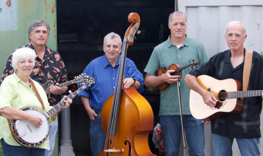 Five adults stand holding musical instruments, including a banjo, mandolin, upright bass, violin, and guitar, in front of a metal storage container and green door, smiling at the camera.