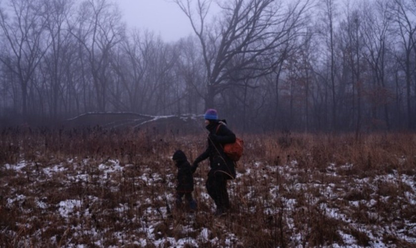 An adult and a child holding hands walk through a snowy, grassy field at dusk. Bare trees and a foggy, overcast sky create a somber winter atmosphere. The adult wears a blue hat and carries an orange backpack.