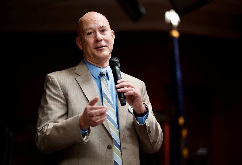 Bald man in a suit speaking into a microphone, gesturing with his hand.