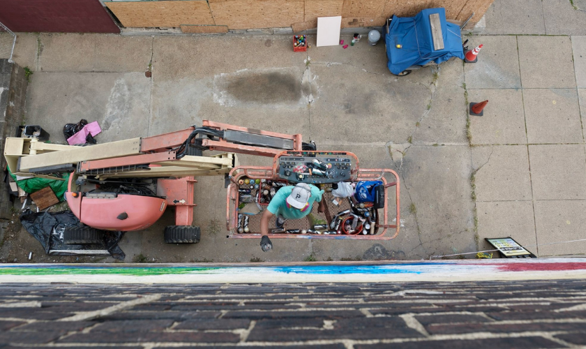 A person in a green shirt and cap stands in a cherry picker lift, working next to a brick building. Tools and supplies fill the lift. The scene is viewed from above, with pavement, trash bags, and a blue recycling bin below.