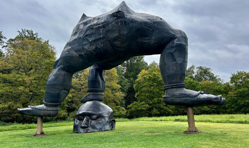 A large outdoor sculpture of a pair of bent legs standing on an upside-down human head, set on a grassy lawn with trees and a cloudy sky in the background.