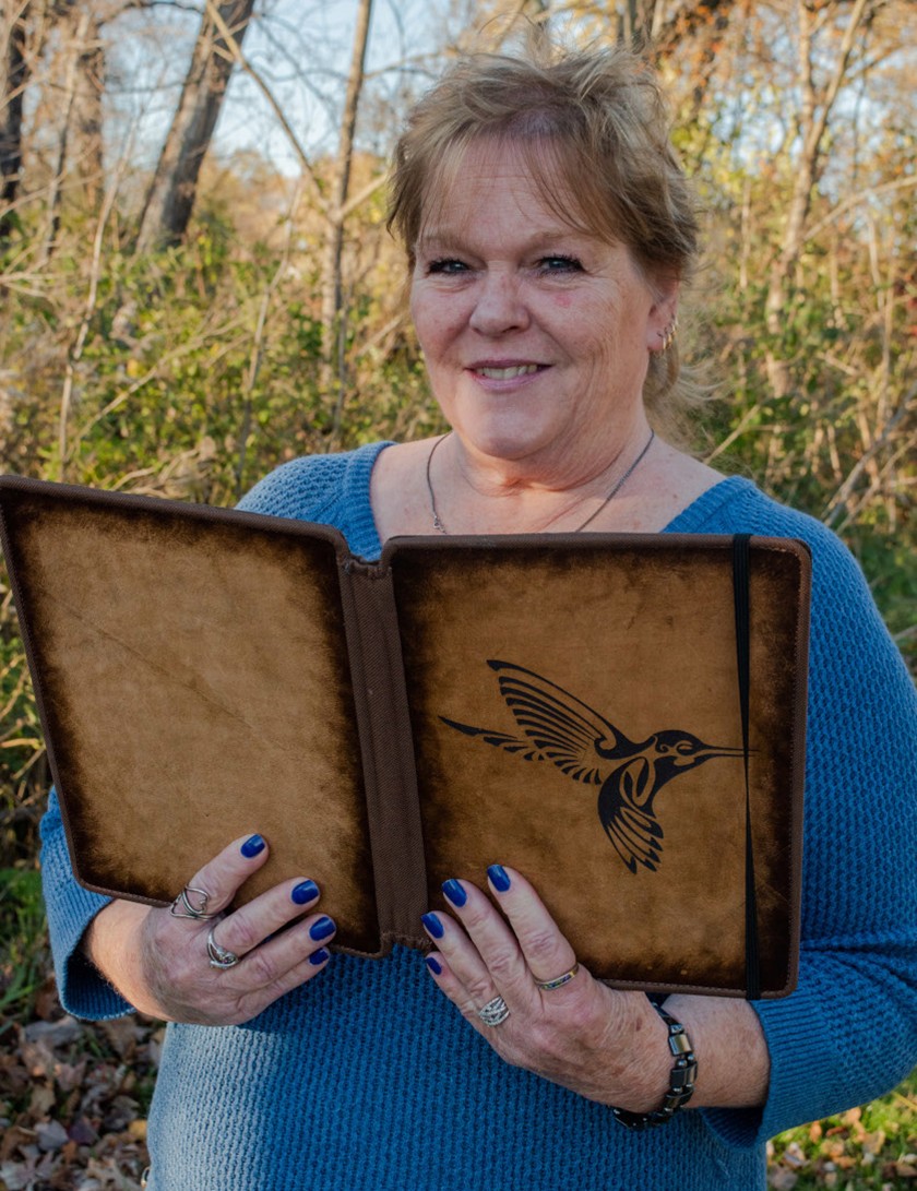 A white woman with pulled-back blonde hair, a necklace, a blue long-sleeved shirt, and blue painted fingernails, smiles while holding a book that has a bird painted on the front cover