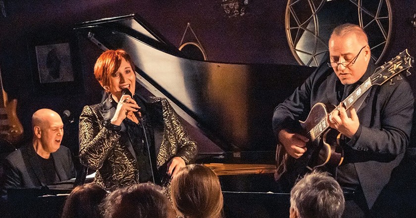 A woman sings in a mic in front of a person playing the piano and beside a person playing electric guitar