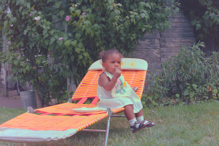 A young child wearing a light dress sits on an orange-striped lounge chair outdoors, eating something with their hand. Lush green bushes and a weathered wooden fence are in the background.