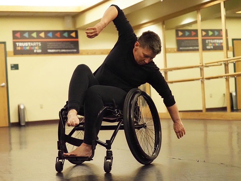 White woman with short hair, black long sleeved shirt, and black leggings dances in a wheelchair
