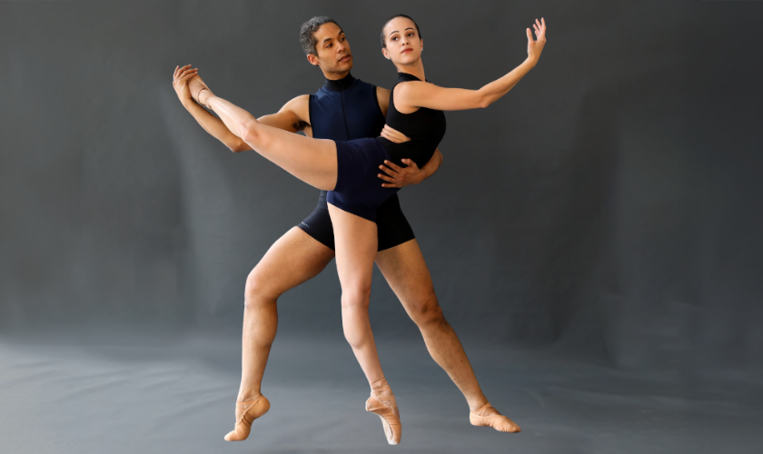 Two ballet dancers in black outfits perform a pose; one lifts the other, who extends her leg and arms gracefully while they both balance on pointe shoes against a neutral background.