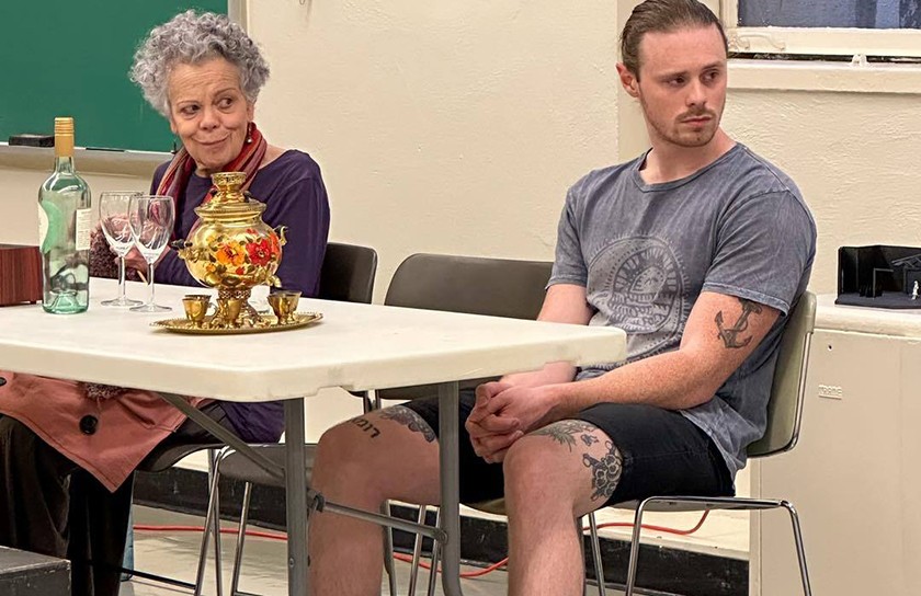 An older woman with gray hair smirks while sitting beside a young white man with brown hair and a goatee