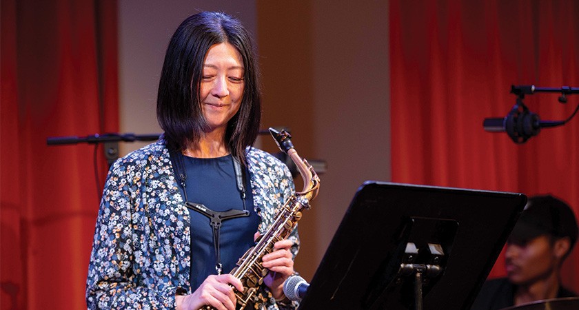 A woman with shoulder-length straight black hair, blue shirt, and a blue patterned blazer, holds a saxophone in front of a music stand