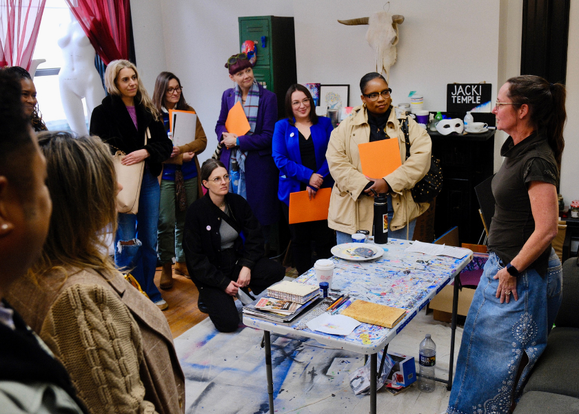 A group of people listens to a woman speak in a colorful art studio