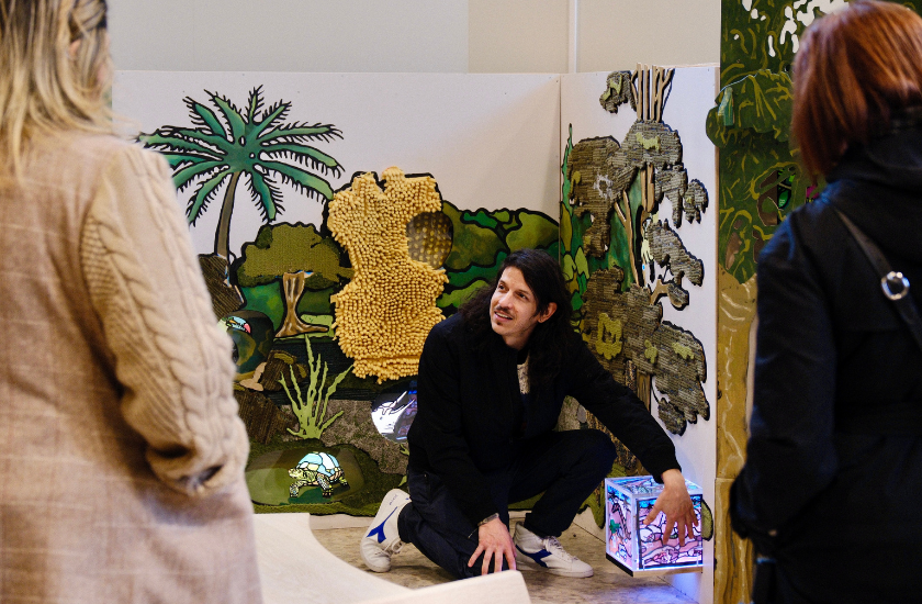 A person crouches by a colorful jungle-themed display, talking to two others
