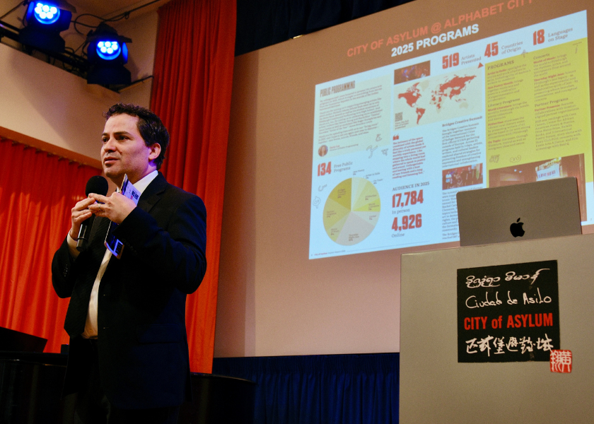 A man speaks onstage beside a projector screen displaying program statistics and charts