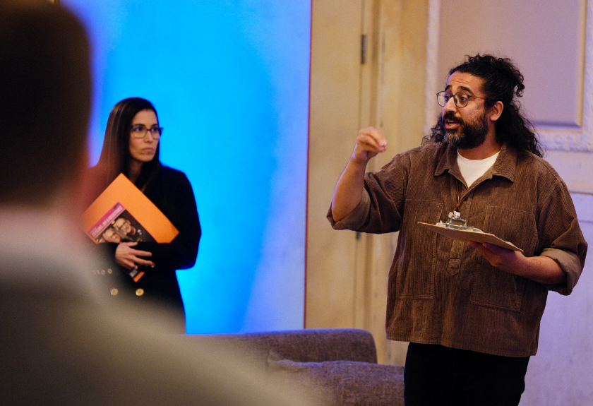 A man speaks while holding a clipboard; a woman stands nearby holding folders.