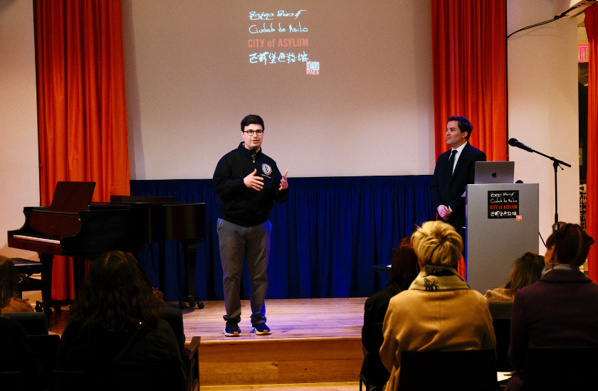 Two men speak on stage to an audience in front of a screen at an indoor event.