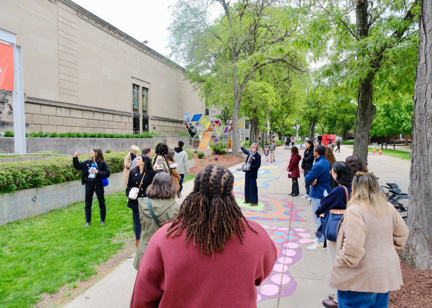 A group of people listen to a guide outdoors near a museum and colorful ground art.