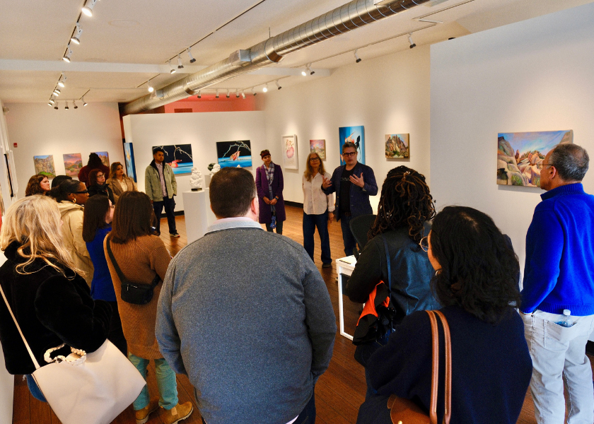 A group listens to a speaker in an art gallery with paintings displayed on white walls.