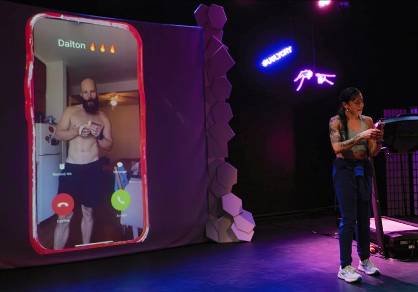 A woman with dark hair, tank top, blue jeans, and sneakers stands on stage looking on her phone while a large image of a phone is shown beside her projected on a screen. The large projected phone image shows a white man with a large beard, shorts, and no shirt staring at the camera. The word "Dalton" and three fire emojis are written at the top of the phone screen.