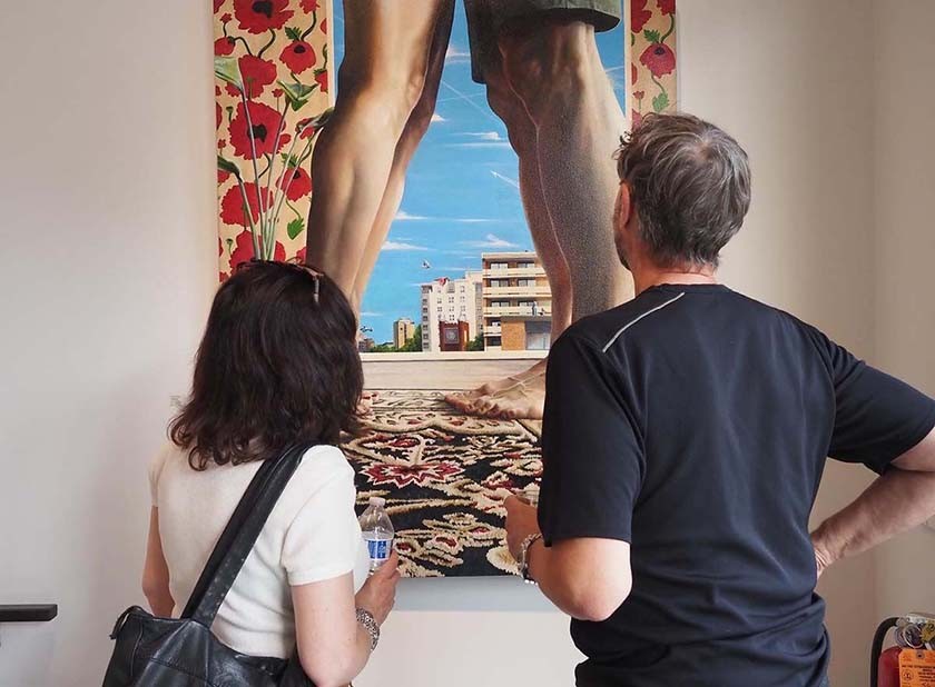 Two people stand with their back to the camera. They're looking at a large multimedia piece hanging on a gallery wall. The artwork shows two sets of bare human legs, buildings, and flowered patterns.