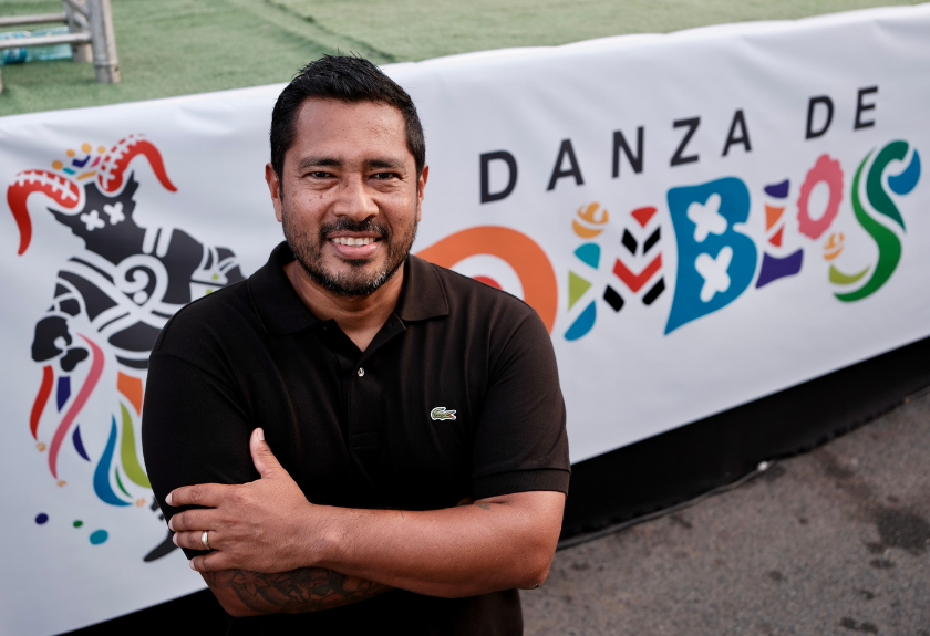 Man in a black polo shirt stands in front of a colorful “Danza de Diablos” banner, smiling with arms crossed.