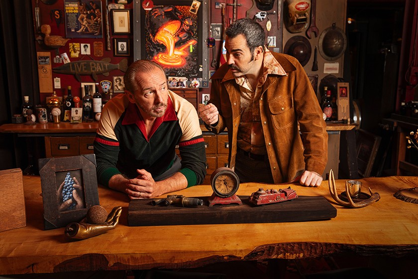 Two white male actors pose behind the counter in a junk shop, surrounded by aged items for sale