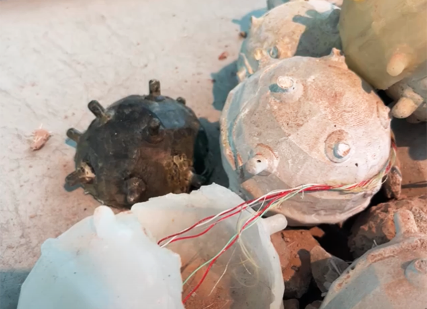 Close-up of several round, spiked objects resembling sea mines on a rough surface. Some mines have exposed wires, and they vary in color, including white, black, and beige.