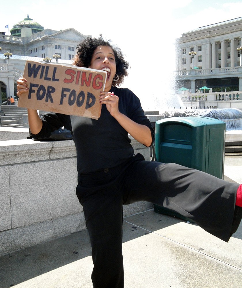 Former GPAC staff Christiane Leach poses with a cardboard sign that says "will sing for food"
