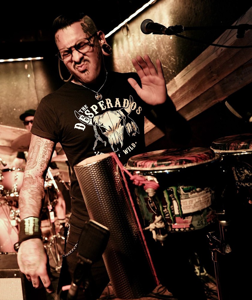 A tattooed musician with gauges plays percussion instruments energetically on stage, wearing a “Desperados” T-shirt. The setting is dimly lit with warm lights, and a drummer is visible in the background.
