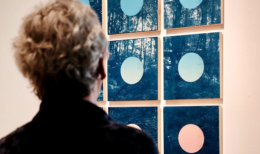A person with curly gray hair views a grid of abstract artworks featuring blue and pink circles over forest imagery displayed on a gallery wall.